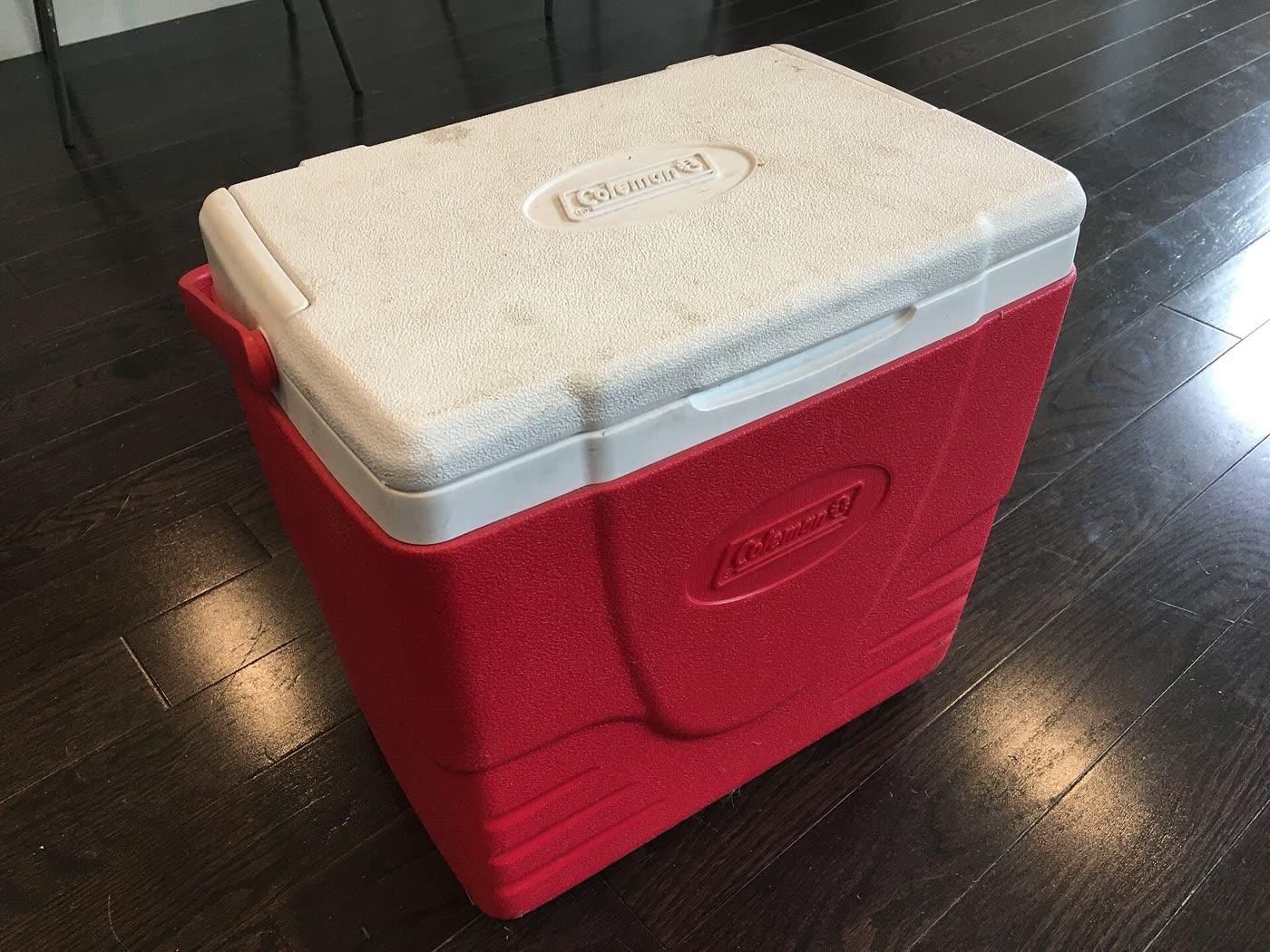 A red Coleman hard cooler with wheels and an extended handle.