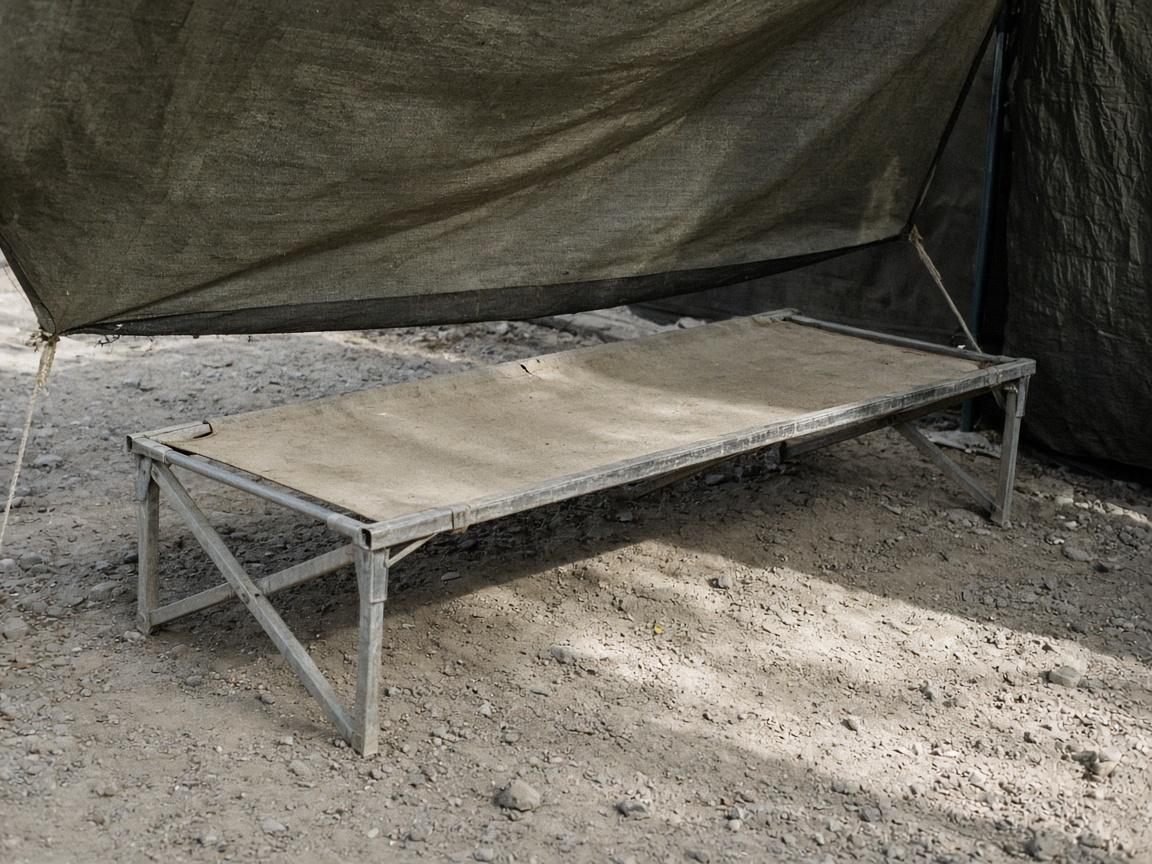 A wide camp cot on pale dust under shade.
