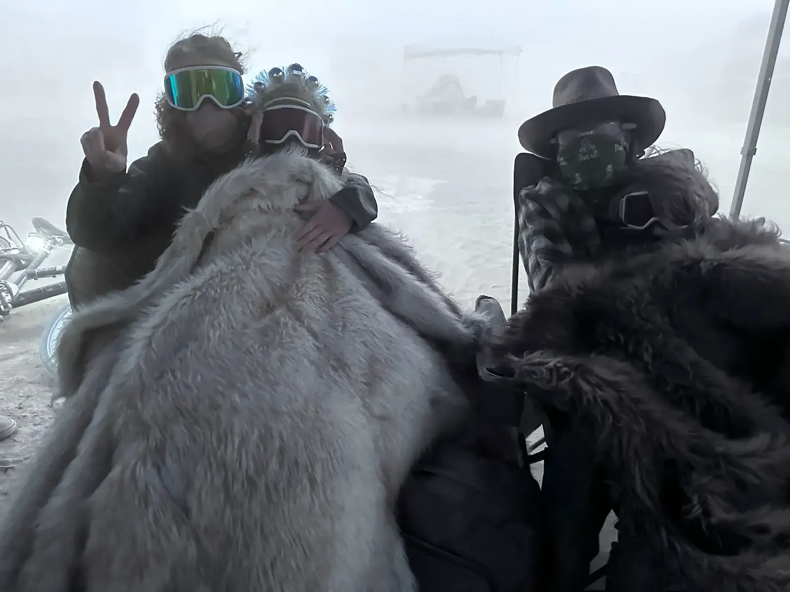 Two people close to the camera in a dust cloud. One wears a fur-lined hood and round goggles. The other wears a cap and ski goggles.