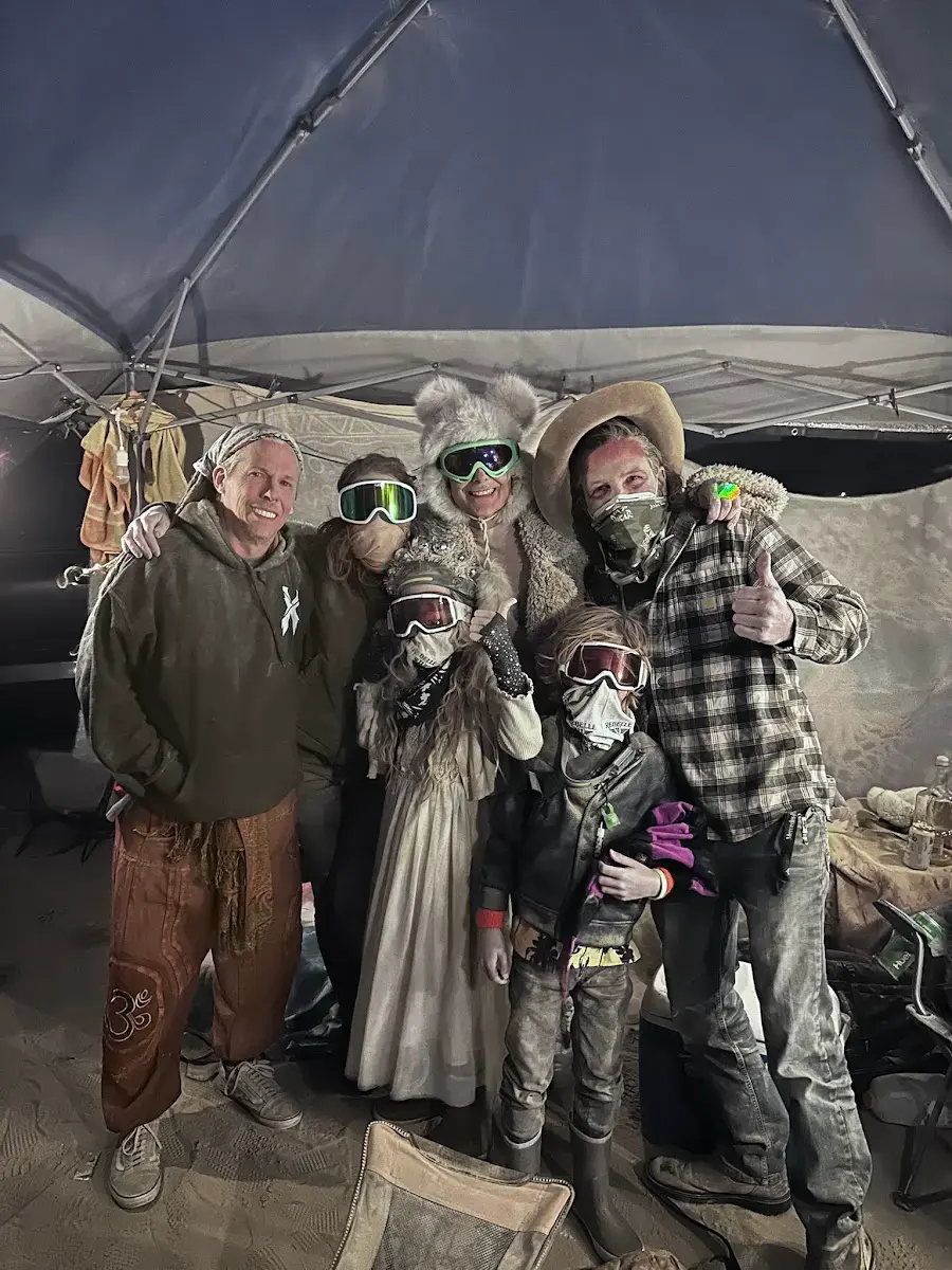 People under a shade structure with a white canopy and patterned fabric walls. One person wears a fur coat and white boots.