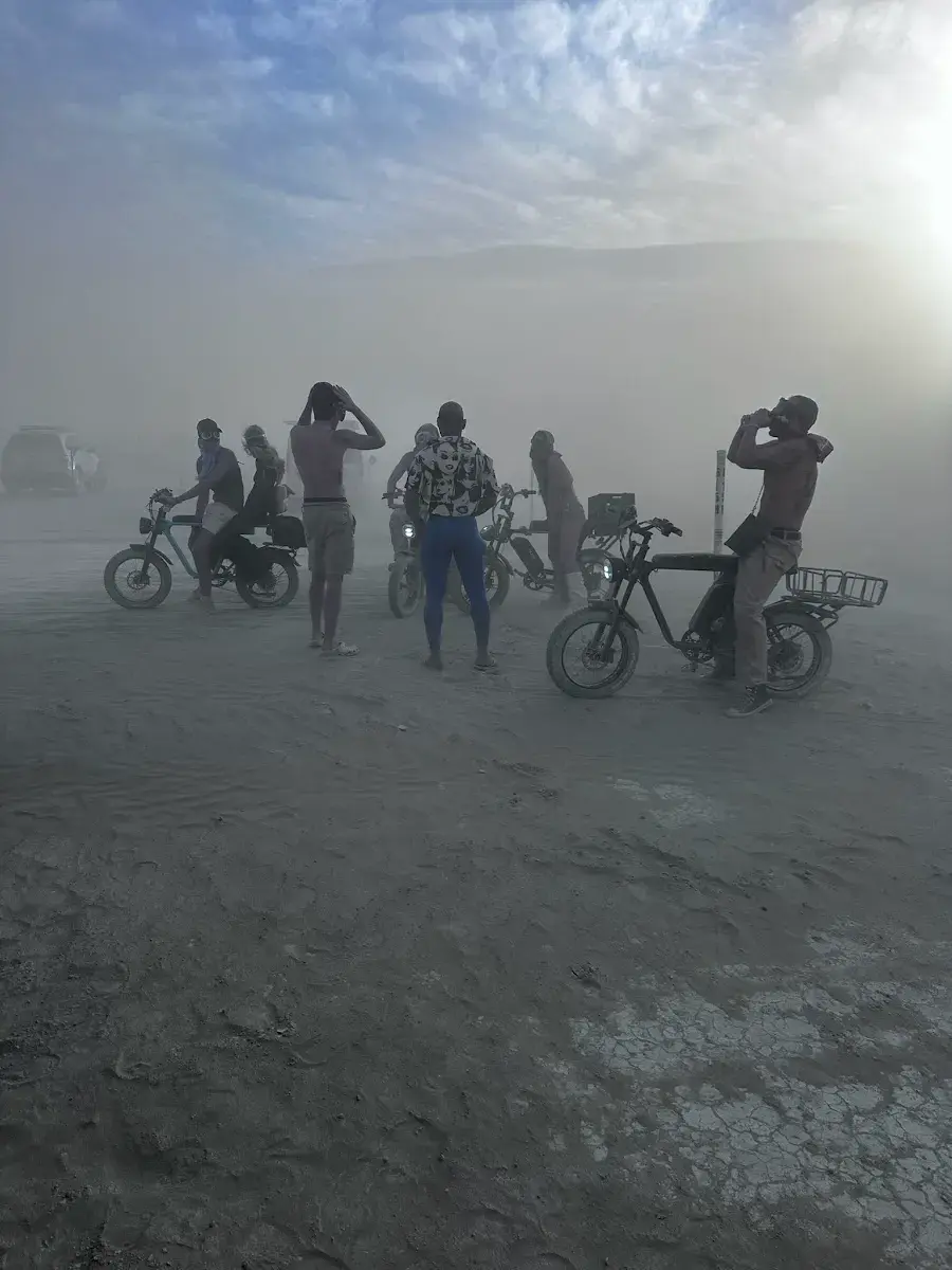 Two people riding bicycles through thick white dust, wearing goggles, with more riders and bicycles behind them.